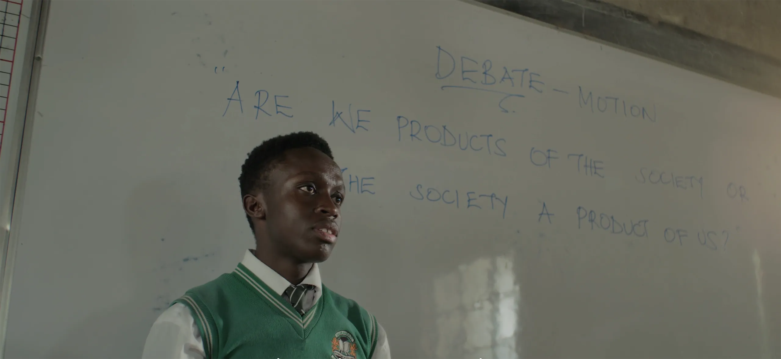 A close up of a young man in a classroom setting with a whiteboard in the background and naturalistic lighting.