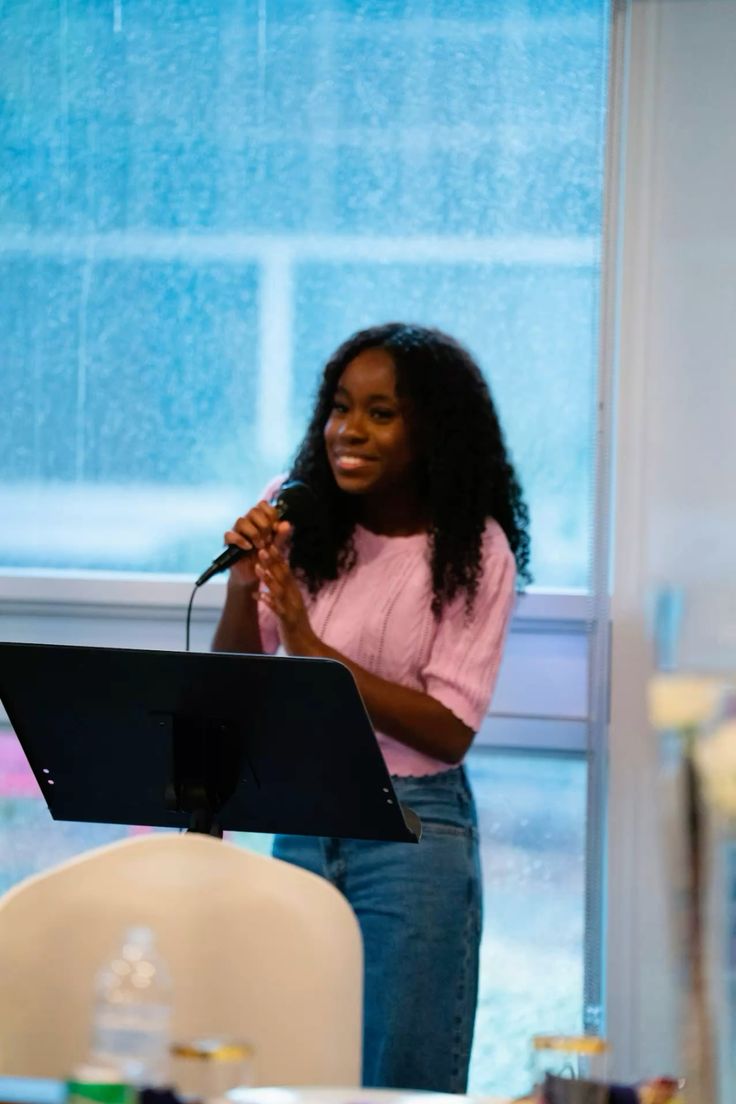 Smiling young woman with long hair standing against a dark green background, holding a finger to her chin.