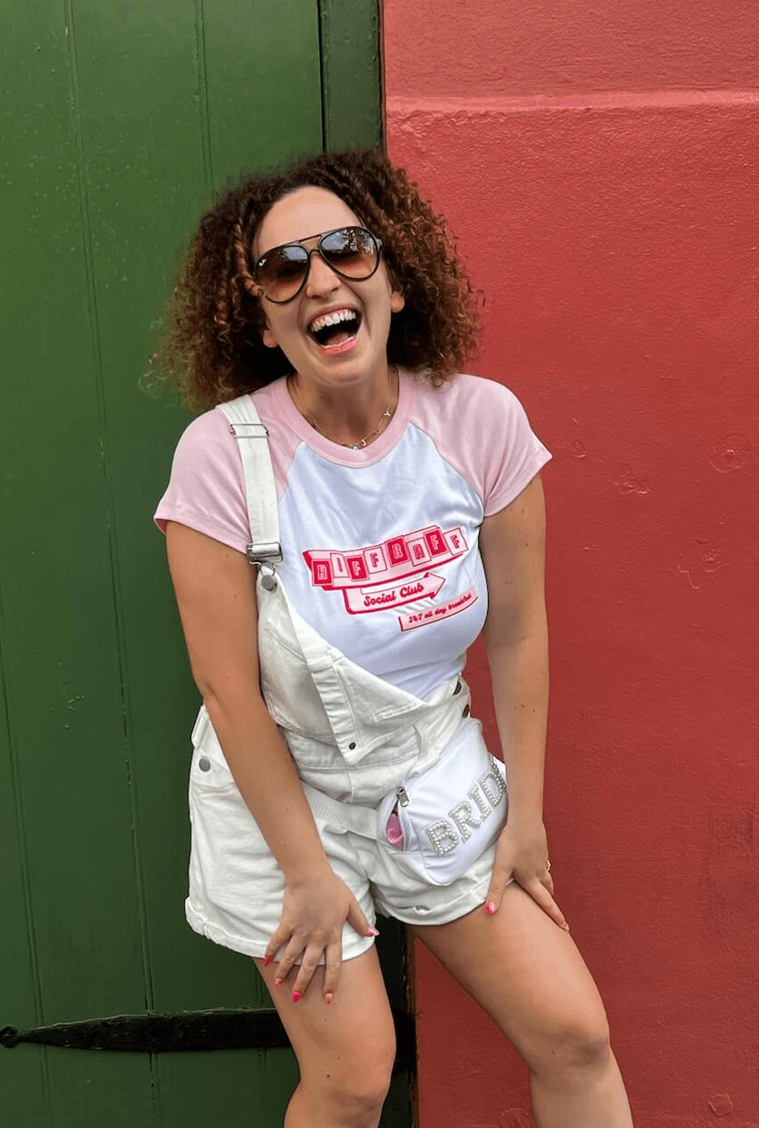 woman with sunglasses and curly dark hair laughing in front of a green and red wall. She is wearing a pink and white baseball baby tee with the riff raff social club diner sign on the front