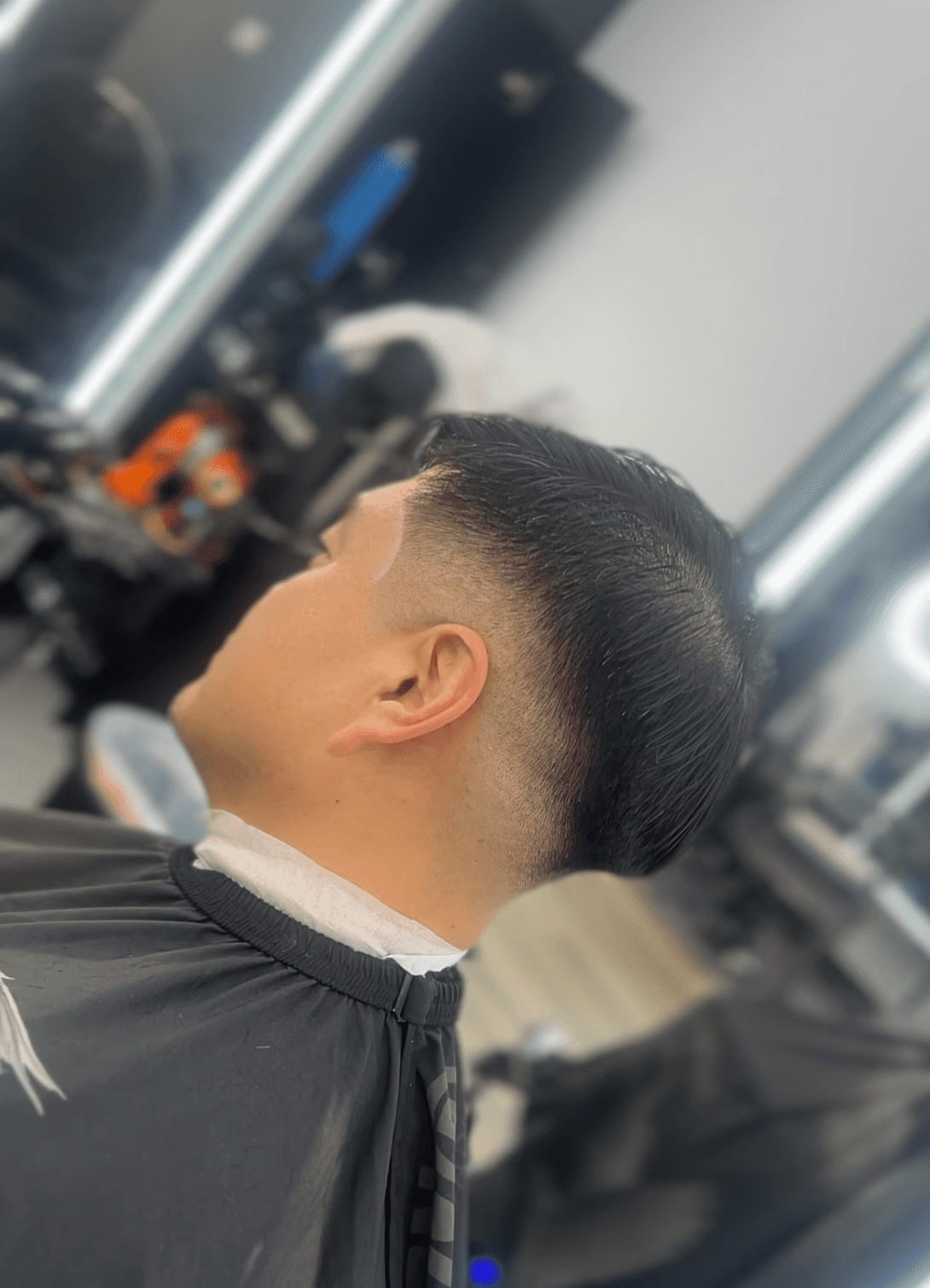Man sitting in a barber chair with a clean fade haircut, short top, and straight fringe while looking at his phone.