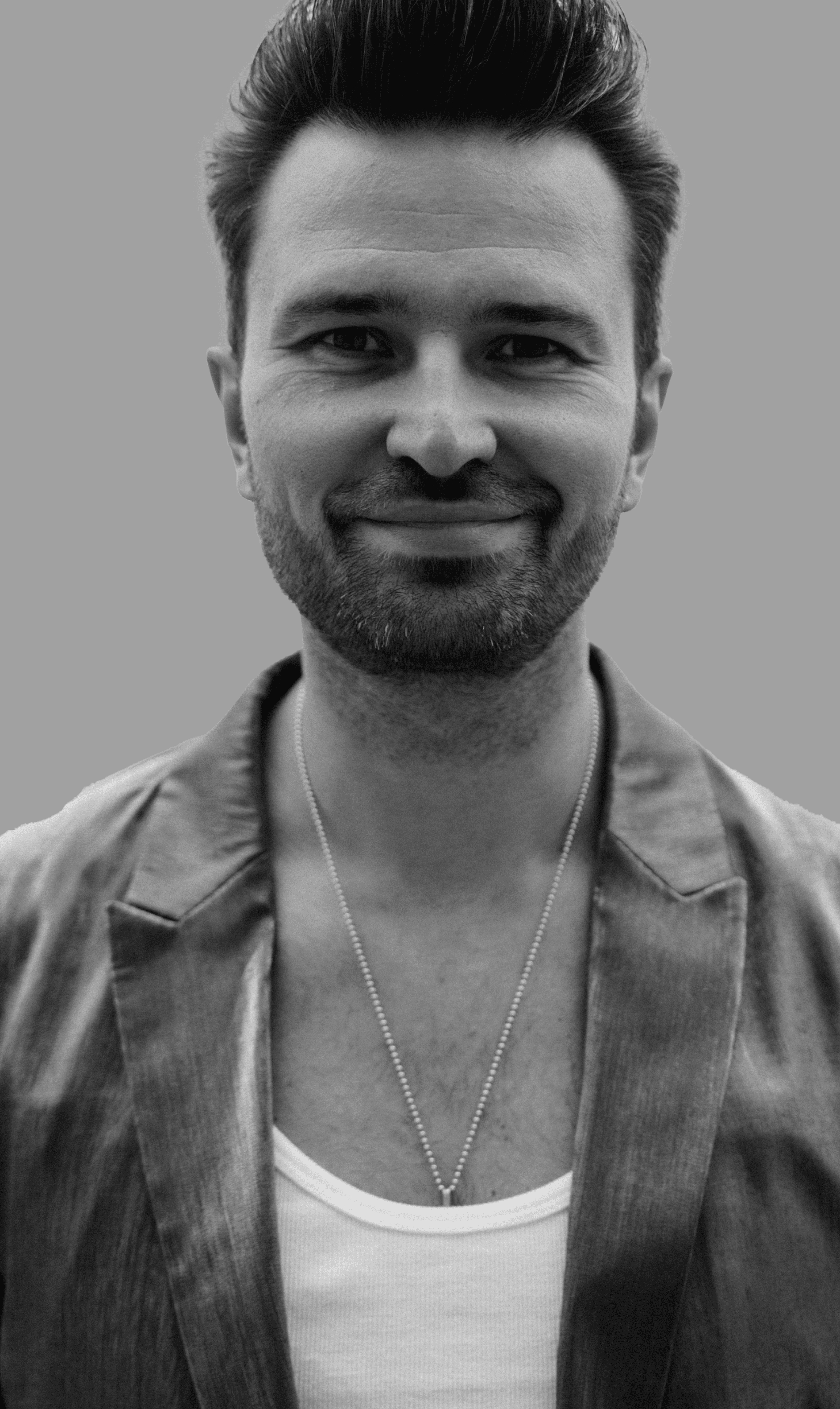 Czarno-biały, bliski portret uśmiechniętego muzyka zespołu Acoustic Shapes w teksturowanej marynarce i białym podkoszulku z delikatnym naszyjnikiem, odzwierciedlający klasę i profesjonalizm.