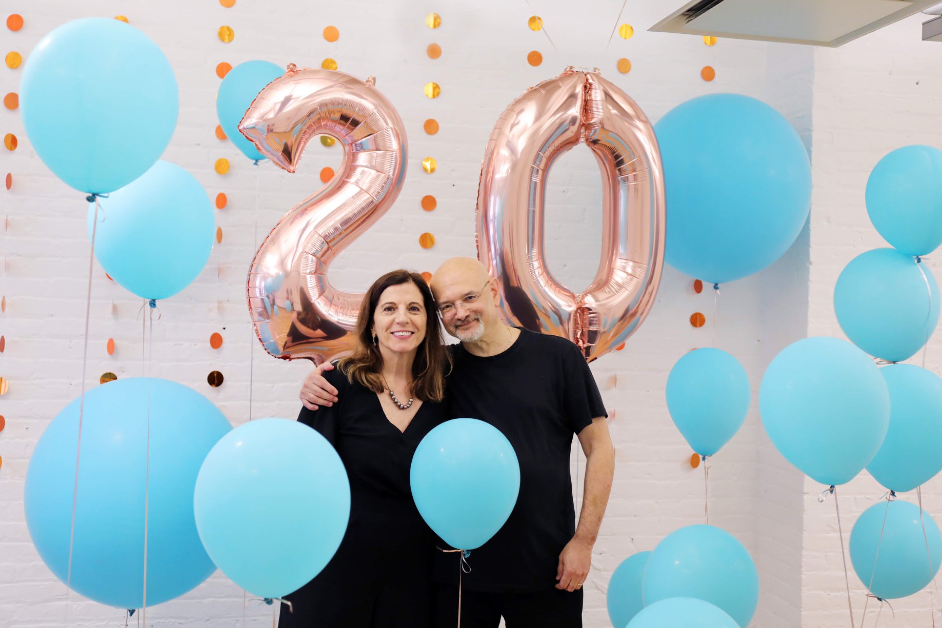 A photo of a man and a woman surrounded by blue balloons and behind them two balloons that resemble the number twenty.