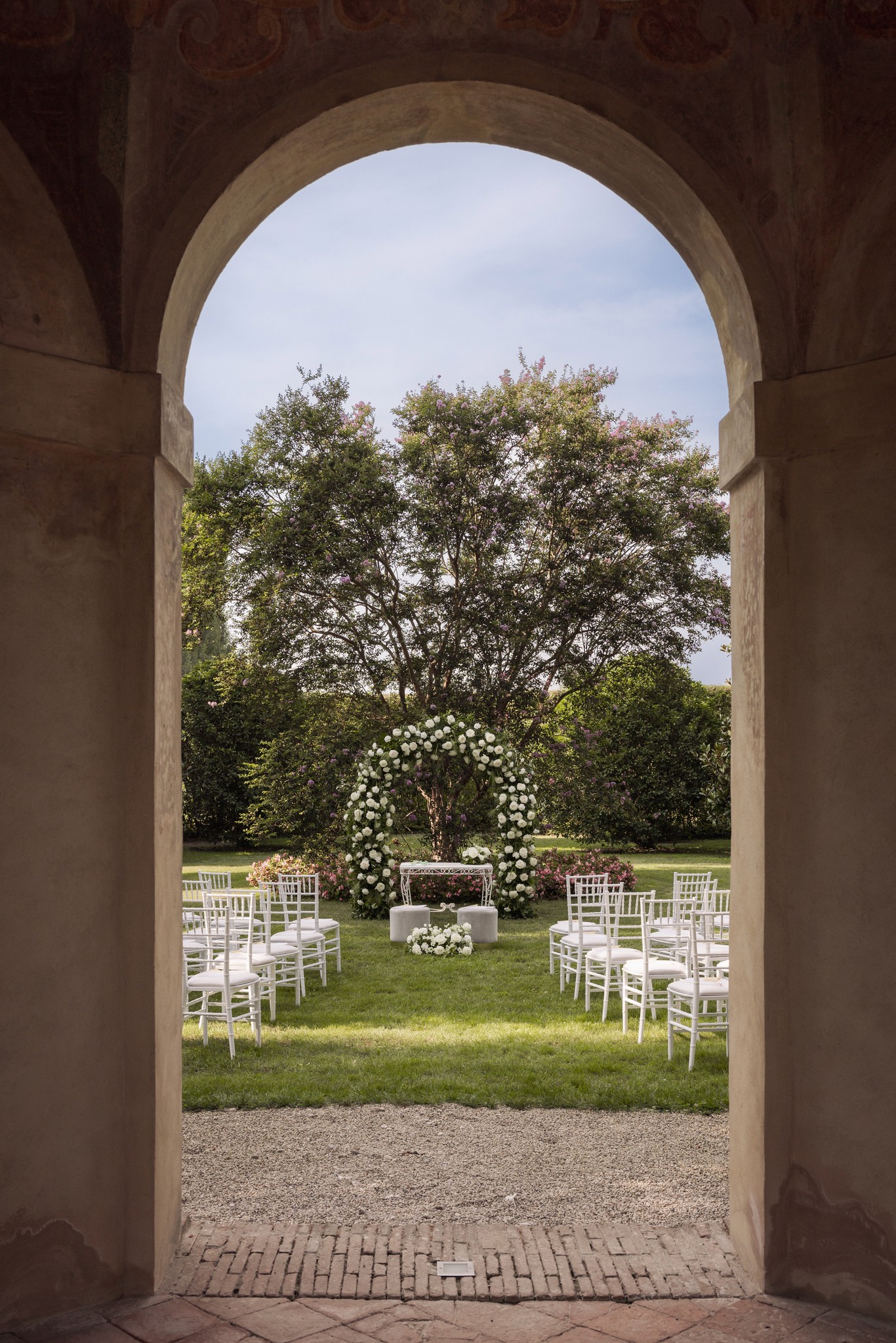 Cerimonia matrimoniale nel giardino di Villa Negri, vista attraverso arco in pietra con allestimento floreale bianco