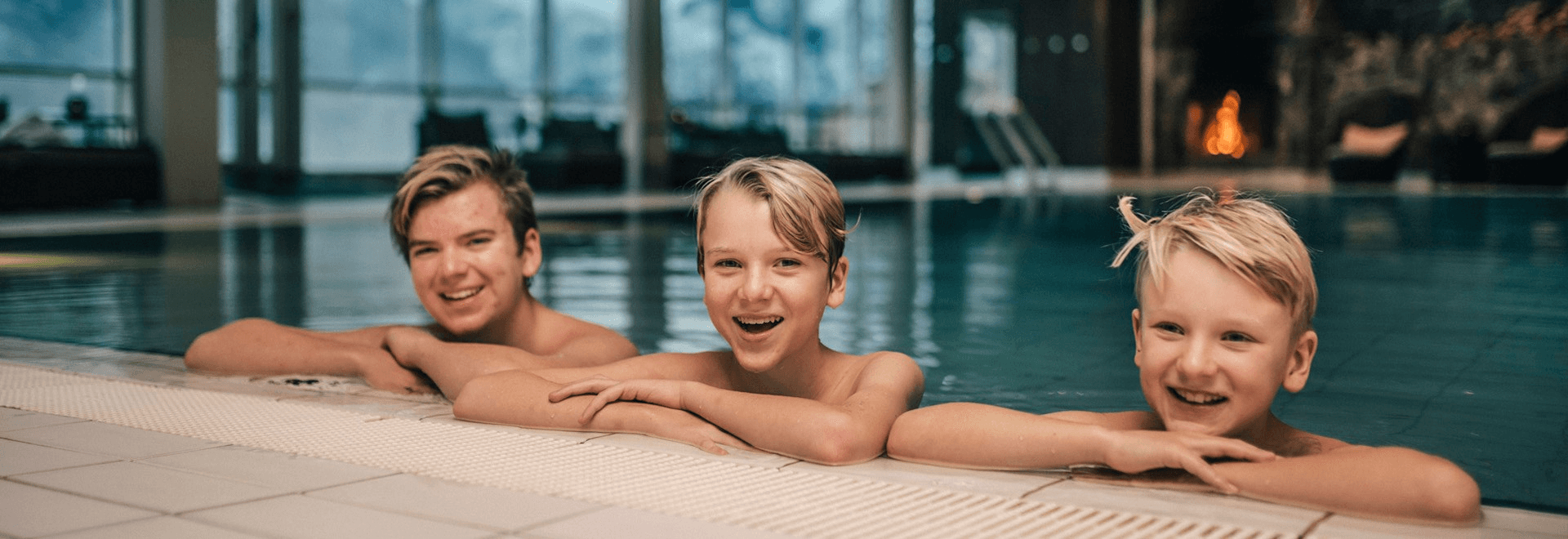 Three children smiling and enjoying a pool, with modern buildings reflecting in the water behind them.