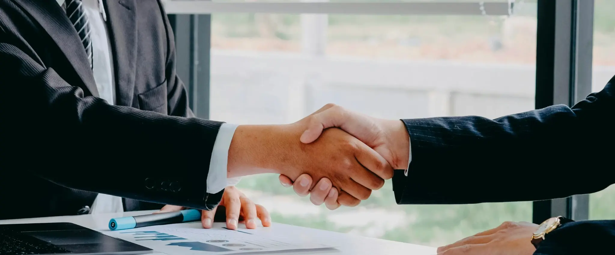 Two people in suits shaking hands over a desk.