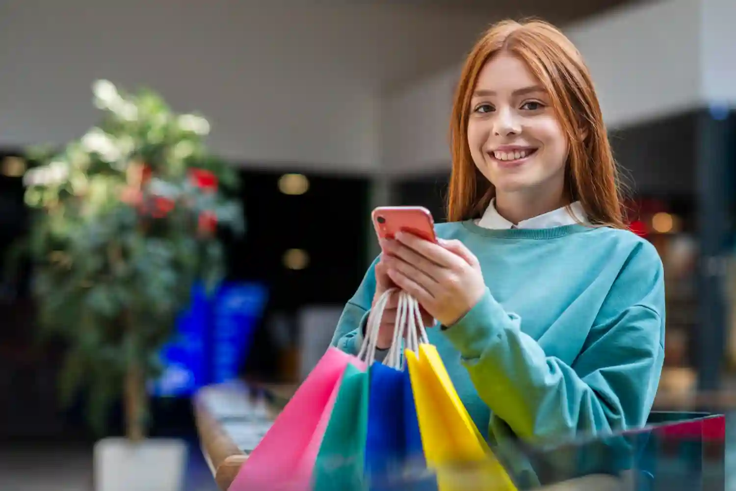Smiling young woman using a mobile app while holding colorful shopping bags in a modern retail mall.