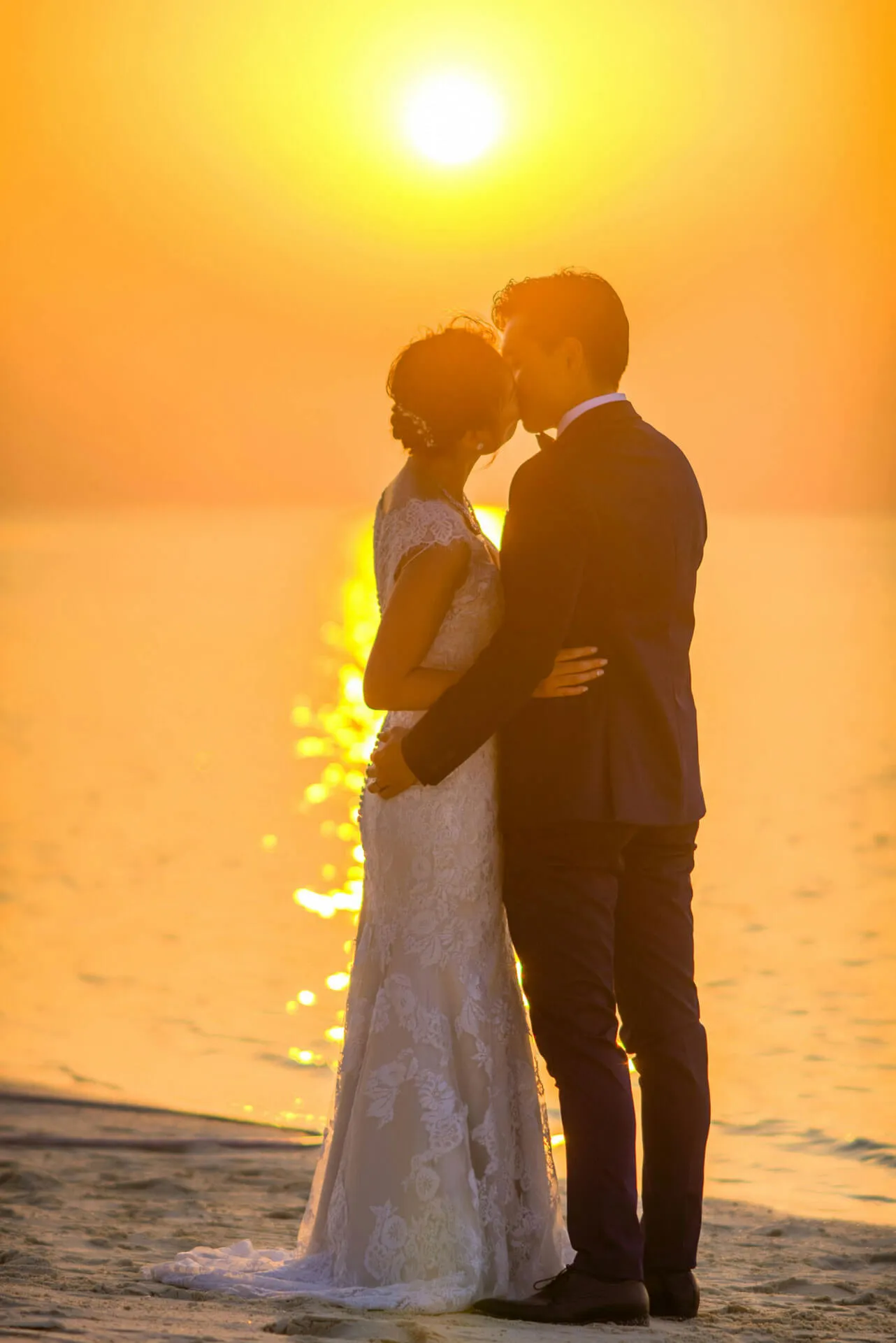 A wedding couple shares a kiss on a tropical Fiji beach at sunset, bathed in warm golden light.