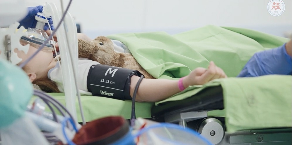 Altatásban fekvő kisgyermek kórházi ágyon, oxigénmaszkkal és plüssjátékkal.