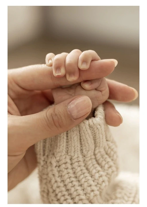 Tiny baby hand holding an adult finger beside a knitted blanket