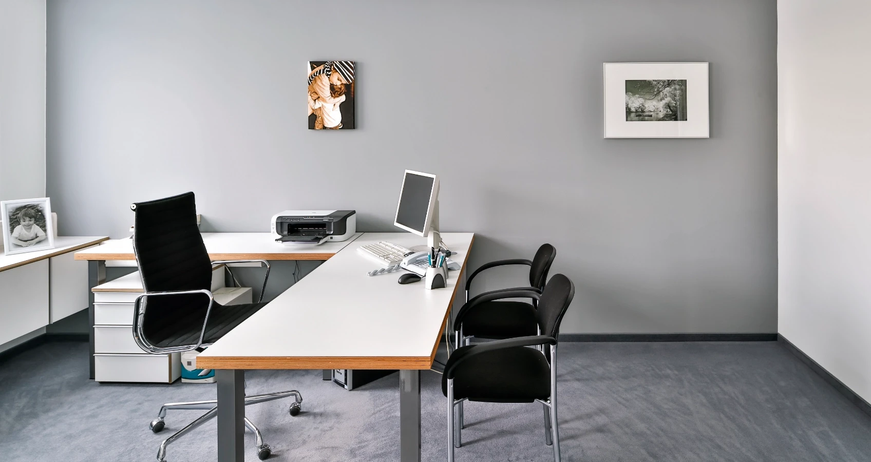 Modernes Büro mit Schreibtisch, Computer, Drucker, Aktenregal und Sitzmöbeln, dekoriert mit gerahmten Bildern.