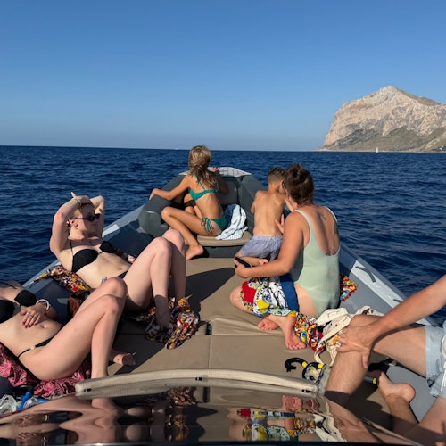 People sunbathing and relaxing on a boat in open water. A rocky coastline is visible in the background under a clear sky.