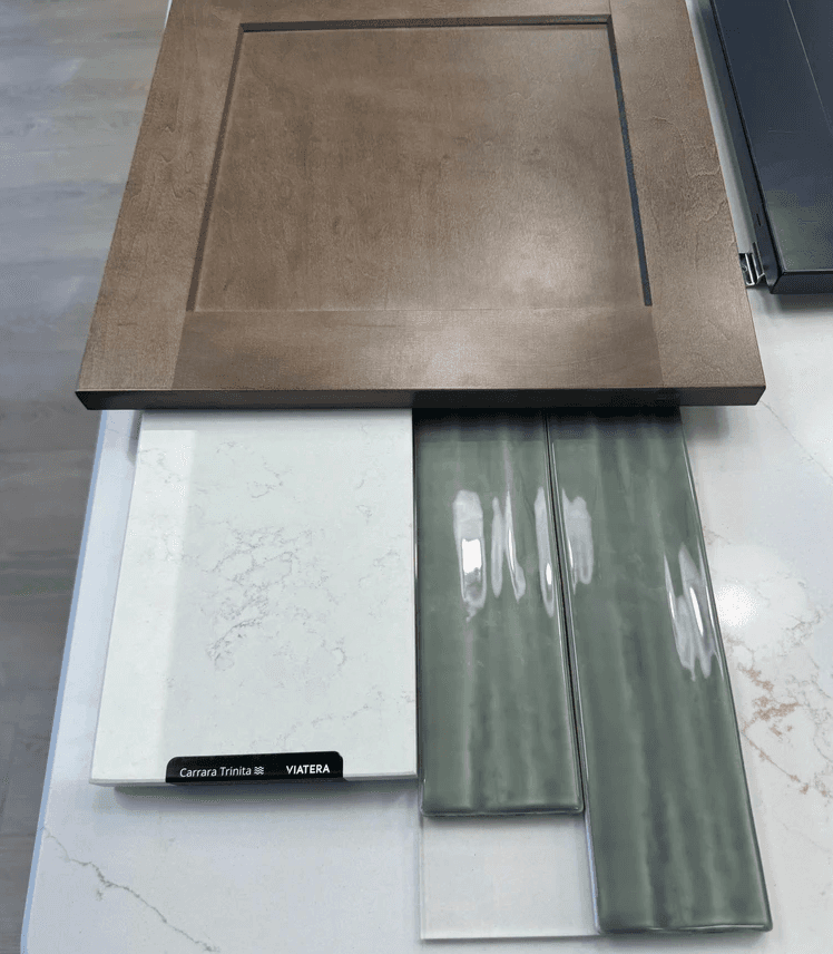 Material sample board showing sage green textured subway tile, taupe shaker cabinet door, and Carrara quartz countertop slab
