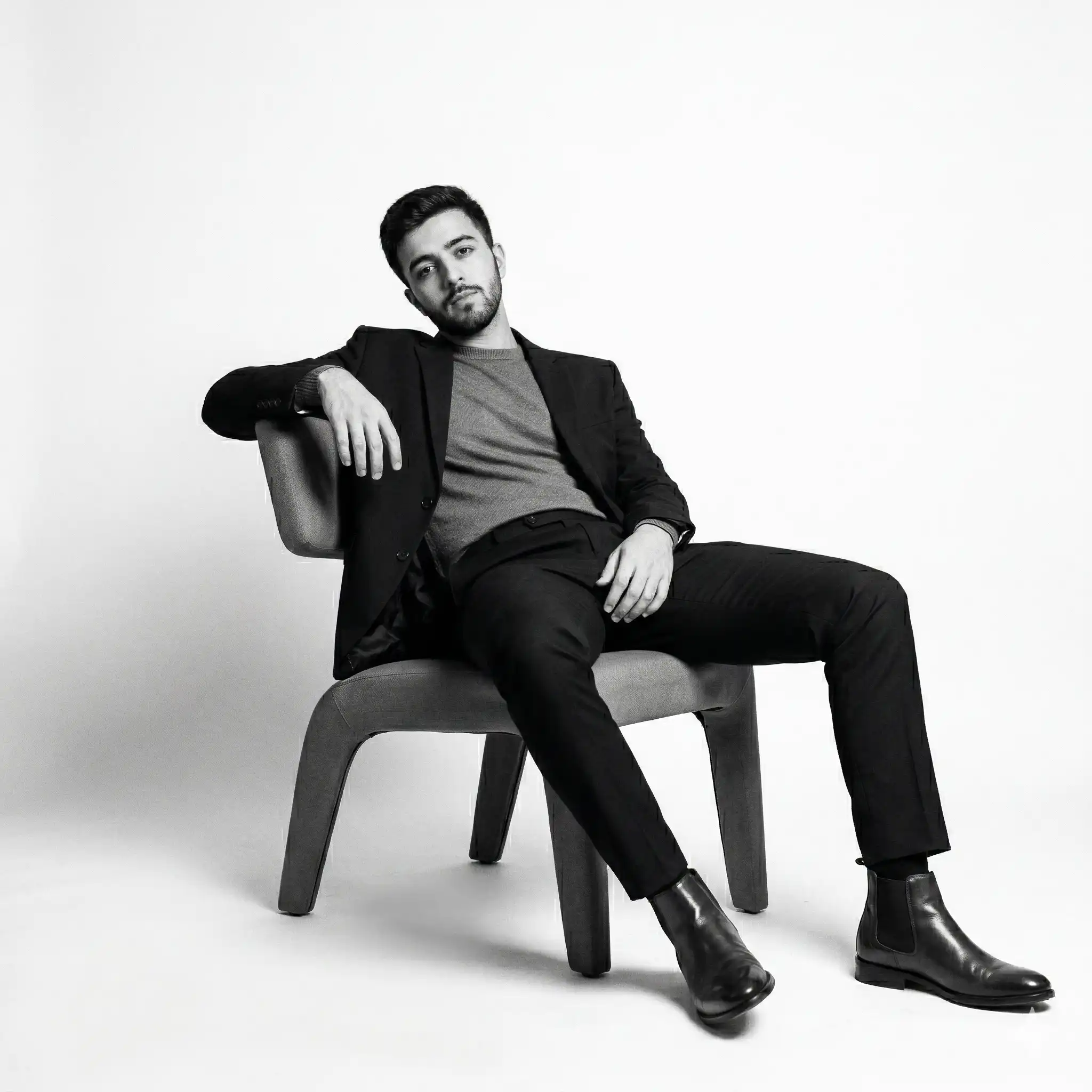 Black and white AI-generated studio portrait of a man sitting in a modern chair wearing a blazer, sweater, and Chelsea boots. Designed for the AI image generation portfolio of Mahendra R.