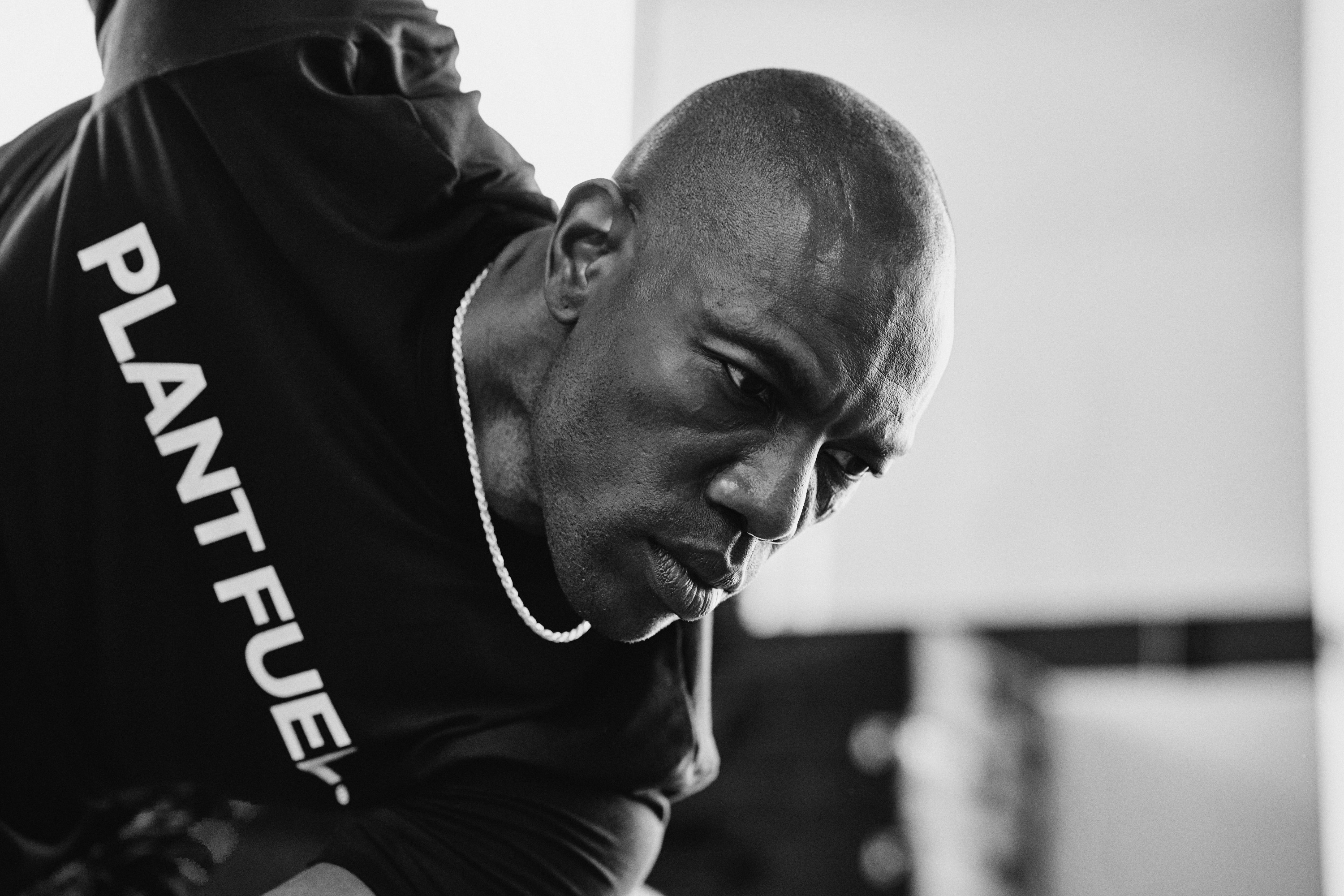 Close-up portrait of a model with pastel pink buzz-cut hair, soft pink eyeshadow, and multiple facial piercings, set against a vibrant green background.