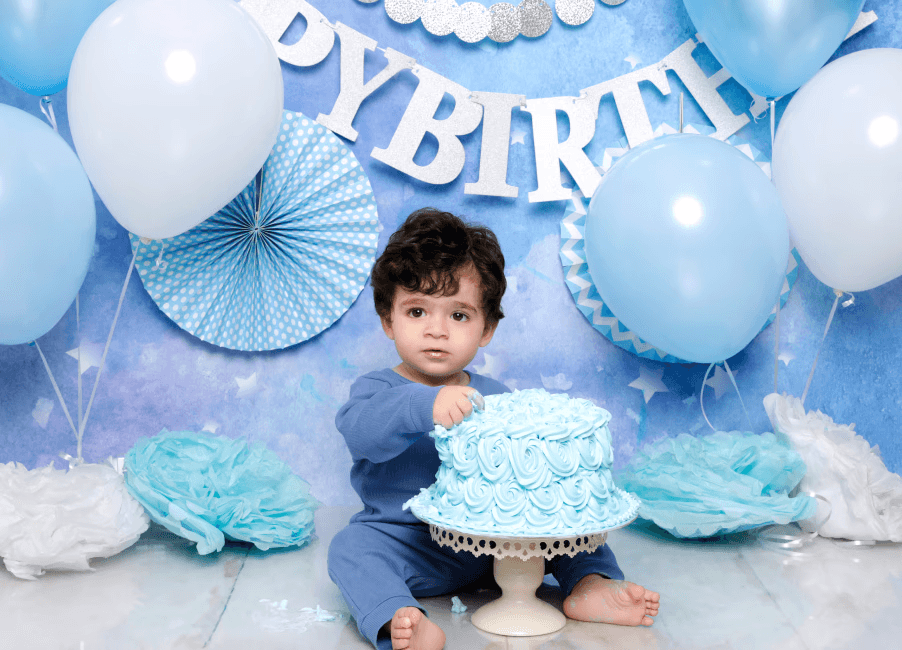 A baby boy is touching a blue floral cake in a blue background, with blue-and-white balloons. There is a “HAPPY BIRTHDAY” banner behind him.