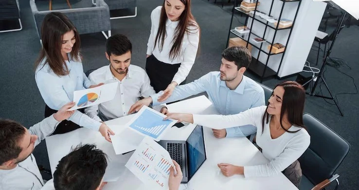 Grupo de pessoas em volta de uma mesa com papéis e laptops, conversando