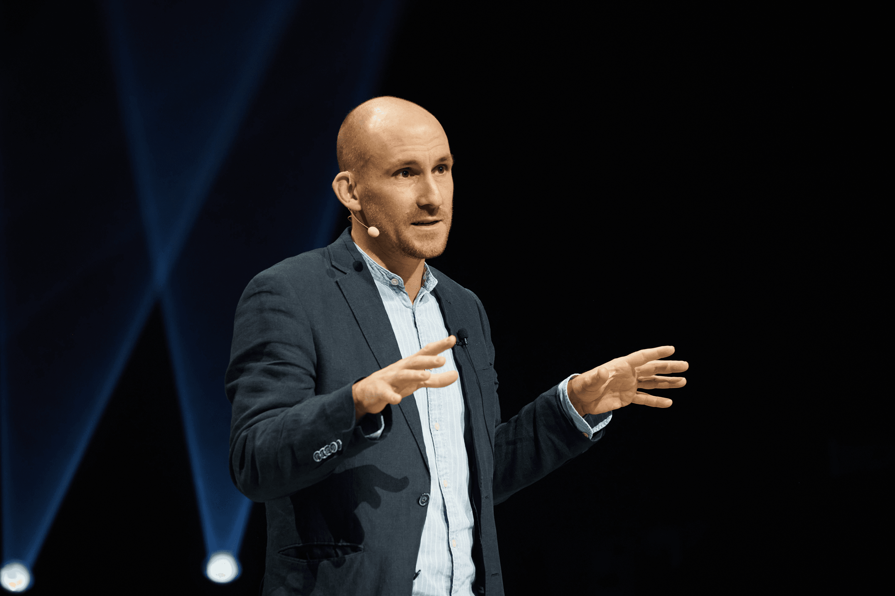 Bald speaker in a dark suit gestures with open hands.