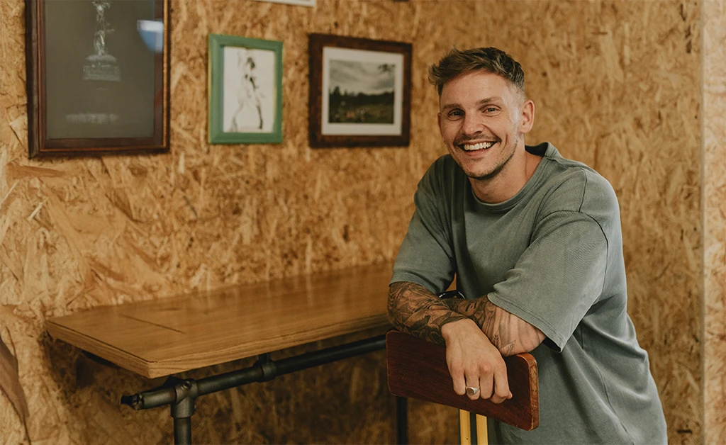 A person wearing headphones sits at a desk with a large curved monitor displaying graphic design software, surrounded by a laptop, smartphone, and potted plant, creating a modern workspace setup.