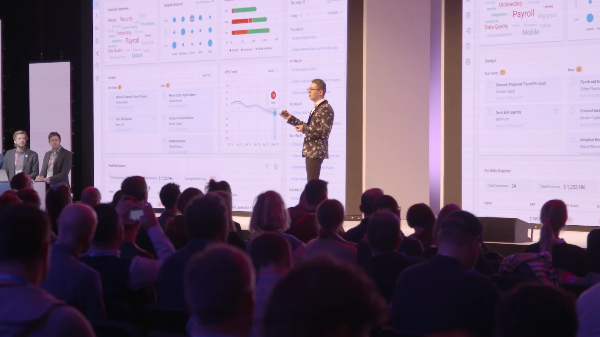 Speaker presenting data and product dashboards on a large screen during a plenary session.