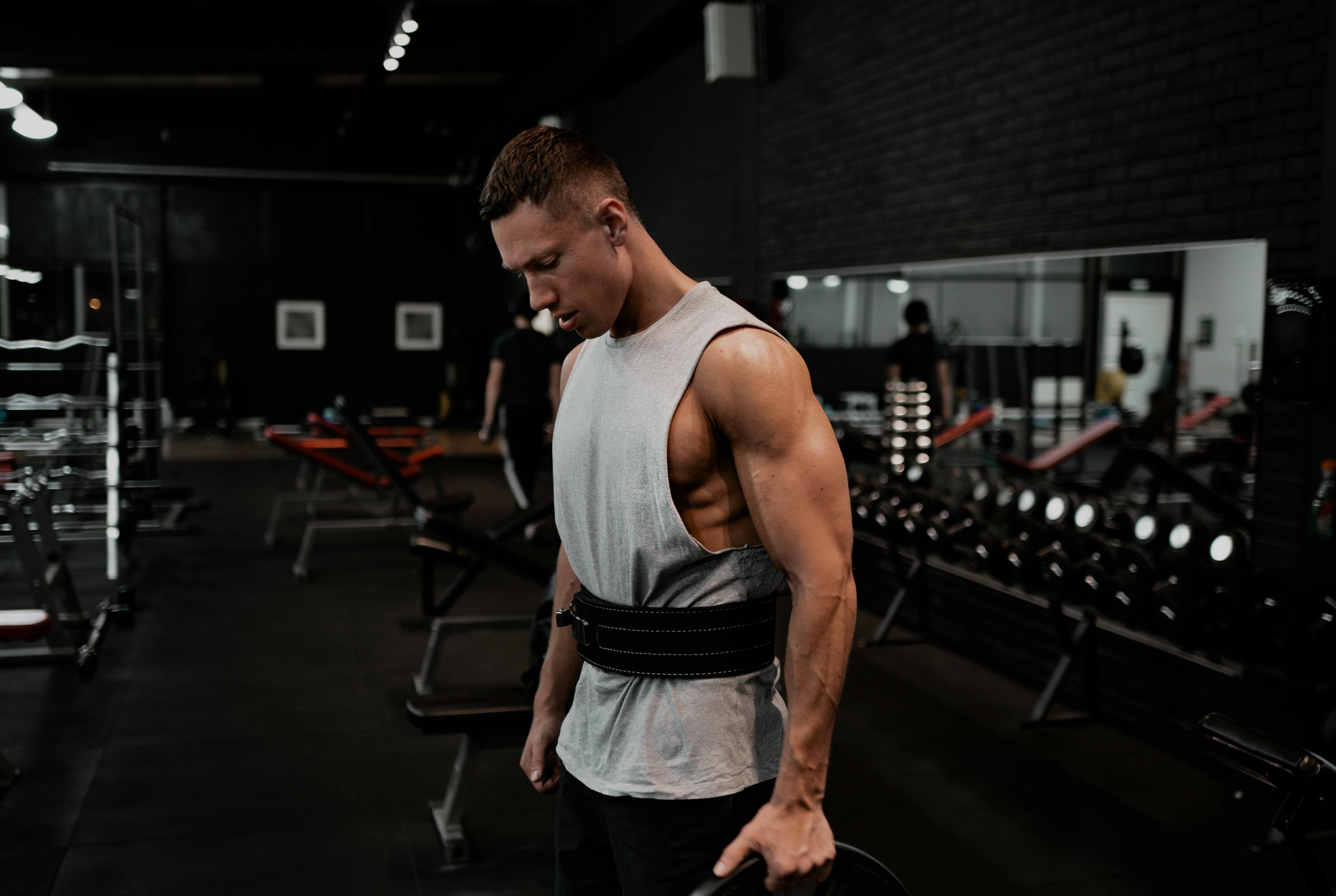 Muscular man standing in a dark gym wearing a weightlifting belt and holding a weight plate between sets.