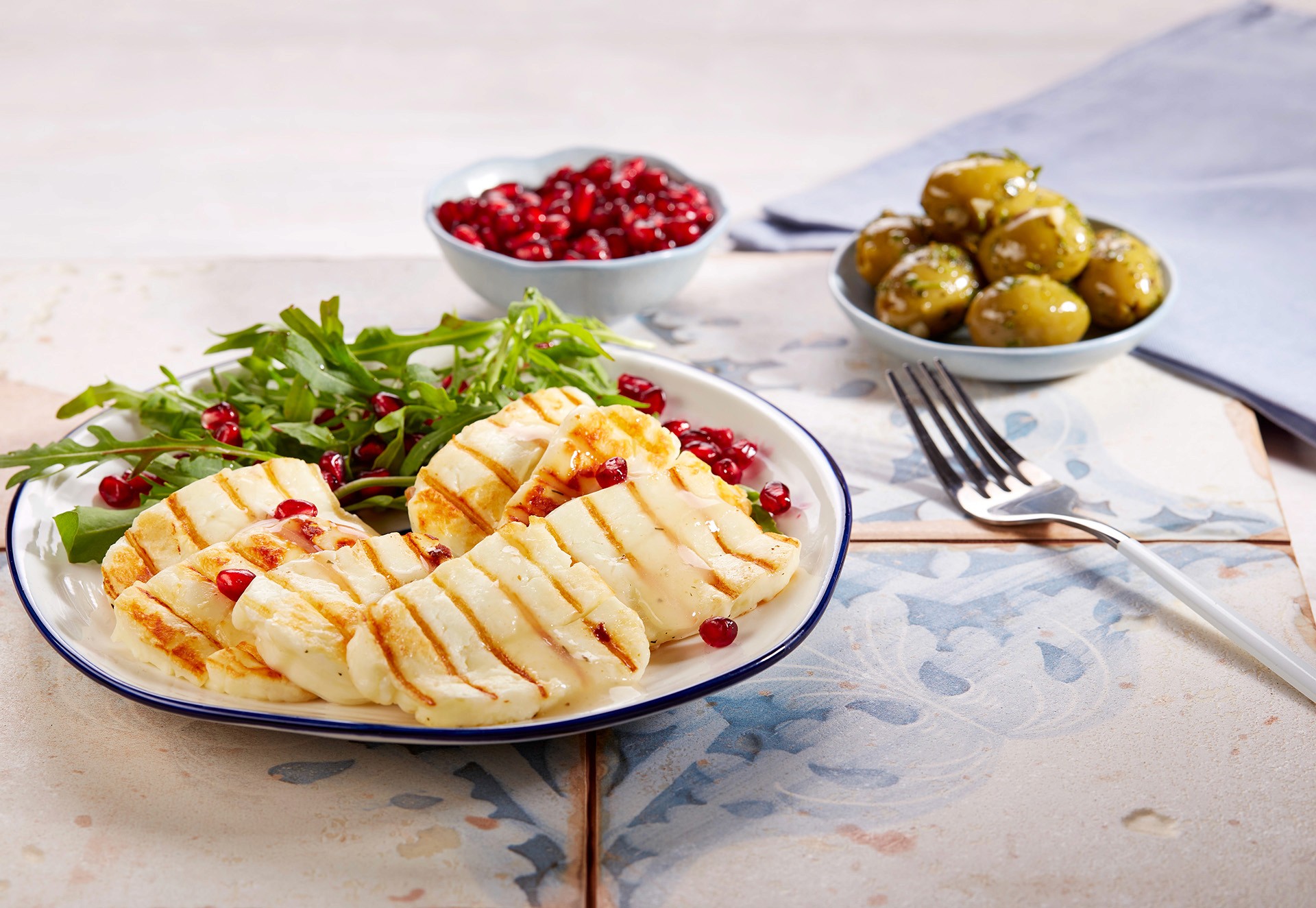 A plate of grilled halloumi cheese garnished with arugula and pomegranate seeds, accompanied by bowls of green olives and fresh pomegranate, set on a decorative tiled table.