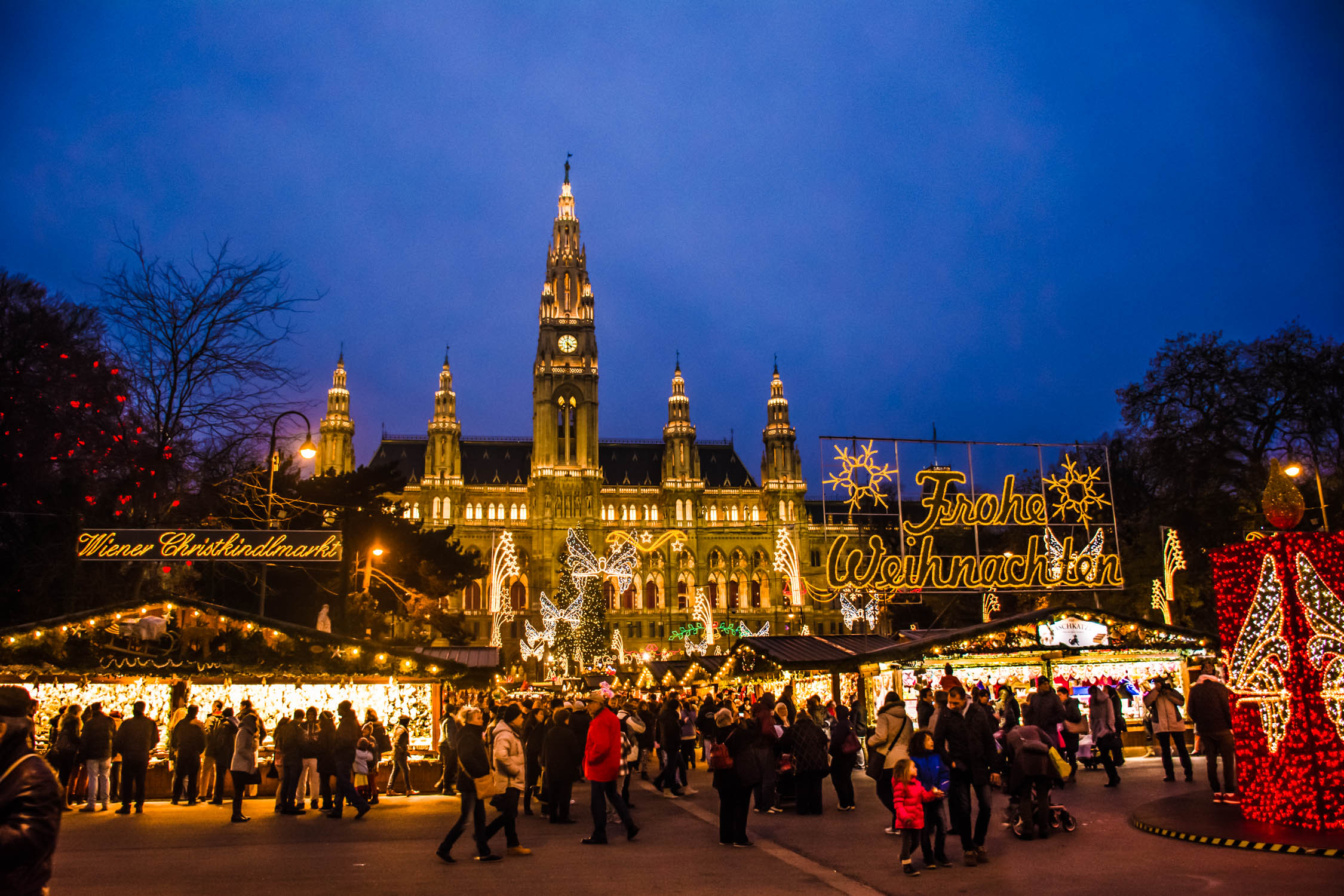 Festive lights illuminate a grand, illuminated building facade and a crowded outdoor market under a deep twilight sky.