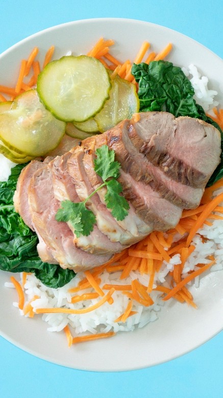 Plate of sliced pork with rice, pickled cucumbers, shredded carrots, sautéed greens, and a side of dipping sauce on a blue background