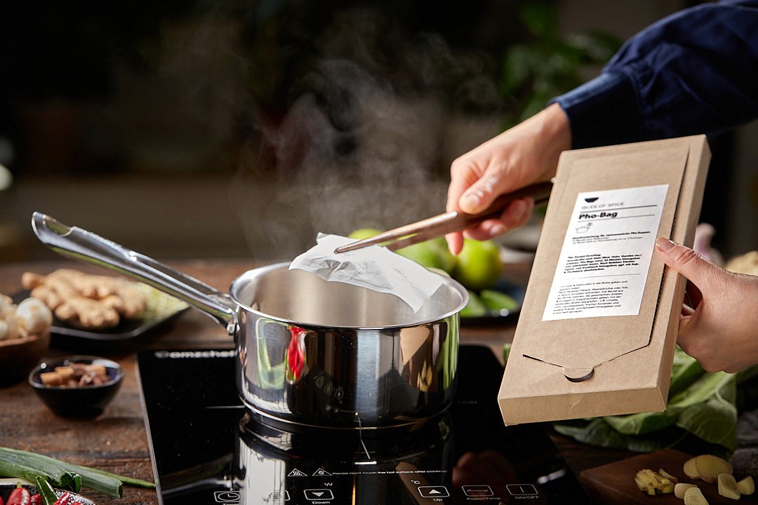 A person adds a Pho Bag to a steaming pot on an induction cooktop, surrounded by fresh ingredients like ginger and leafy greens, creating a vibrant cooking scene.