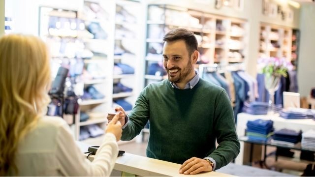 Homem branco de suéter verde entrega um cartão para mulher de cabelos longos e claros que está atrás de um balcão, em uma loja de vestuário