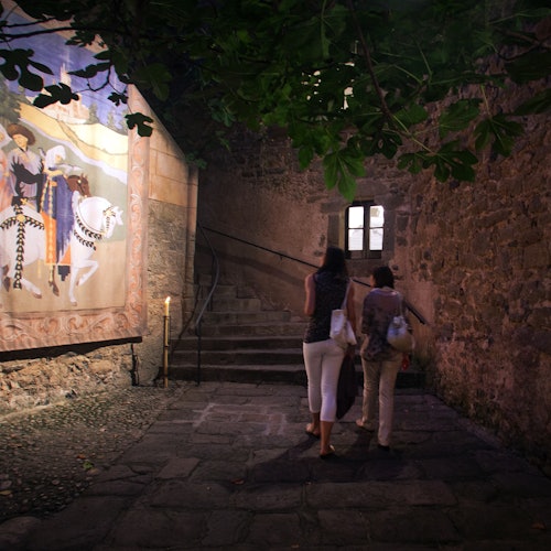 Dalawang babae ang naglalakad paakyat sa isang hagdang bato sa tabi ng isang mural sa pader na naglalarawan ng mga kabayo at mga tao na may kasuotan mula sa kasaysayan.