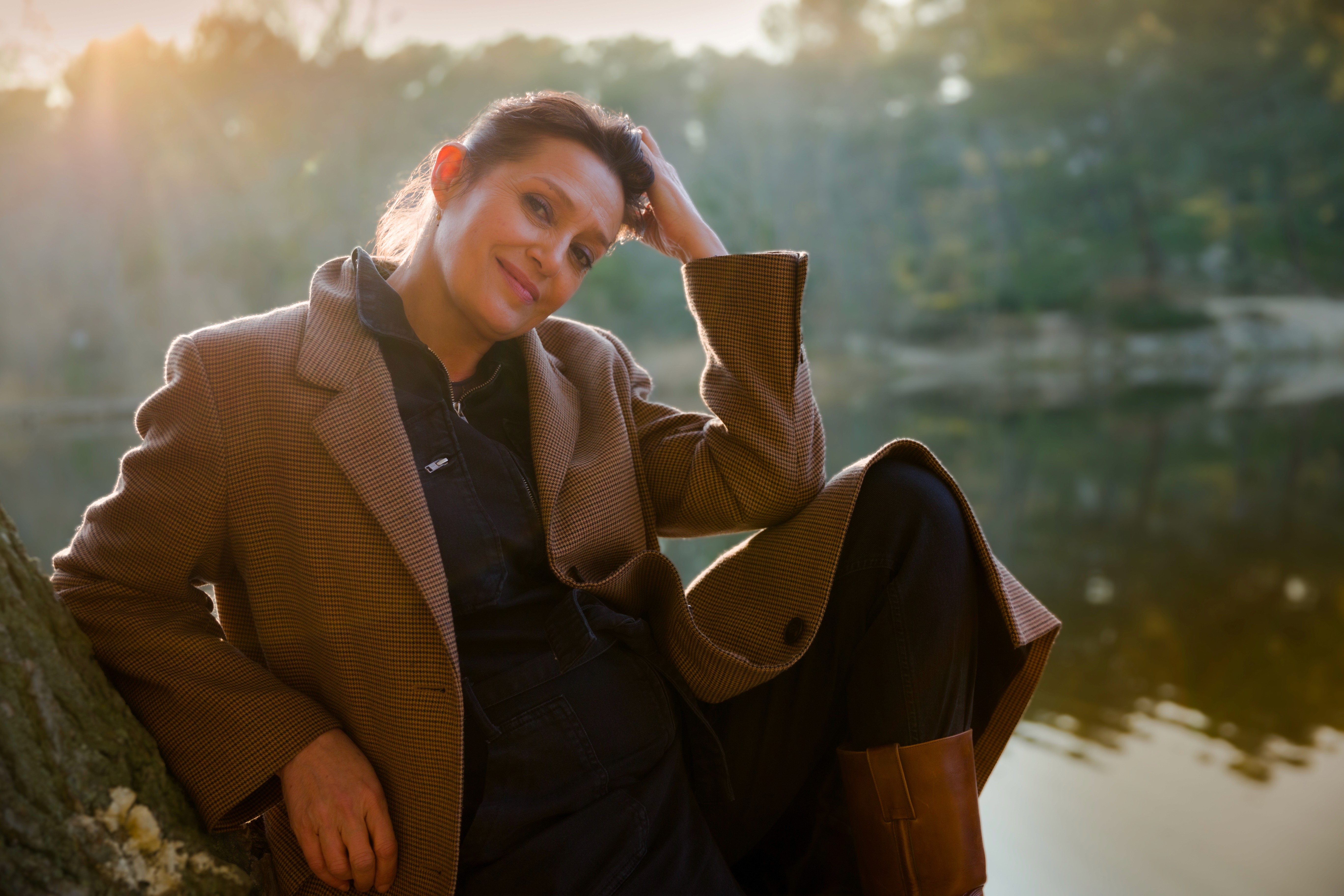 Céline ESCAND, femme élégante se détendant au bord d’un lac au coucher du soleil, portant un manteau marron, ambiance naturelle et apaisante – Le Baume Intégral.