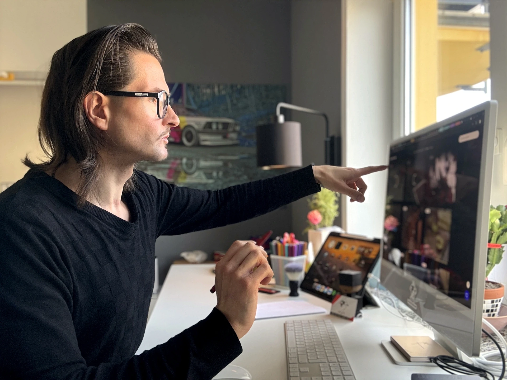 Jens Pfeifer arbeitet fokussiert an einem Apple iMac und zeigt mit dem Finger auf ein Interface-Detail auf dem Bildschirm. Seitliche Perspektive im Studio-Workspace mit iPad, Tastatur, Skizzenblock und Farbstiften auf dem Schreibtisch – visuelle Darstellung eines analytischen Design- und Review-Prozesses in einem professionellen, kreativen Arbeitsumfeld.