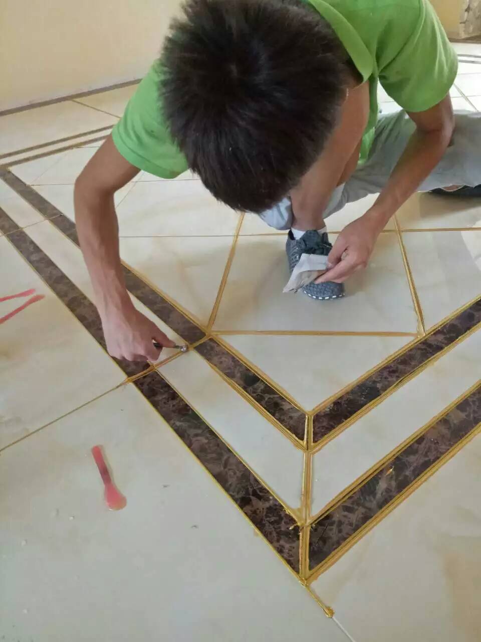 A worker in a green shirt carefully applying gold tile grout along the edges of a tiled floor with a marble inlay. The worker focuses on ensuring precise grout application for a decorative finish.