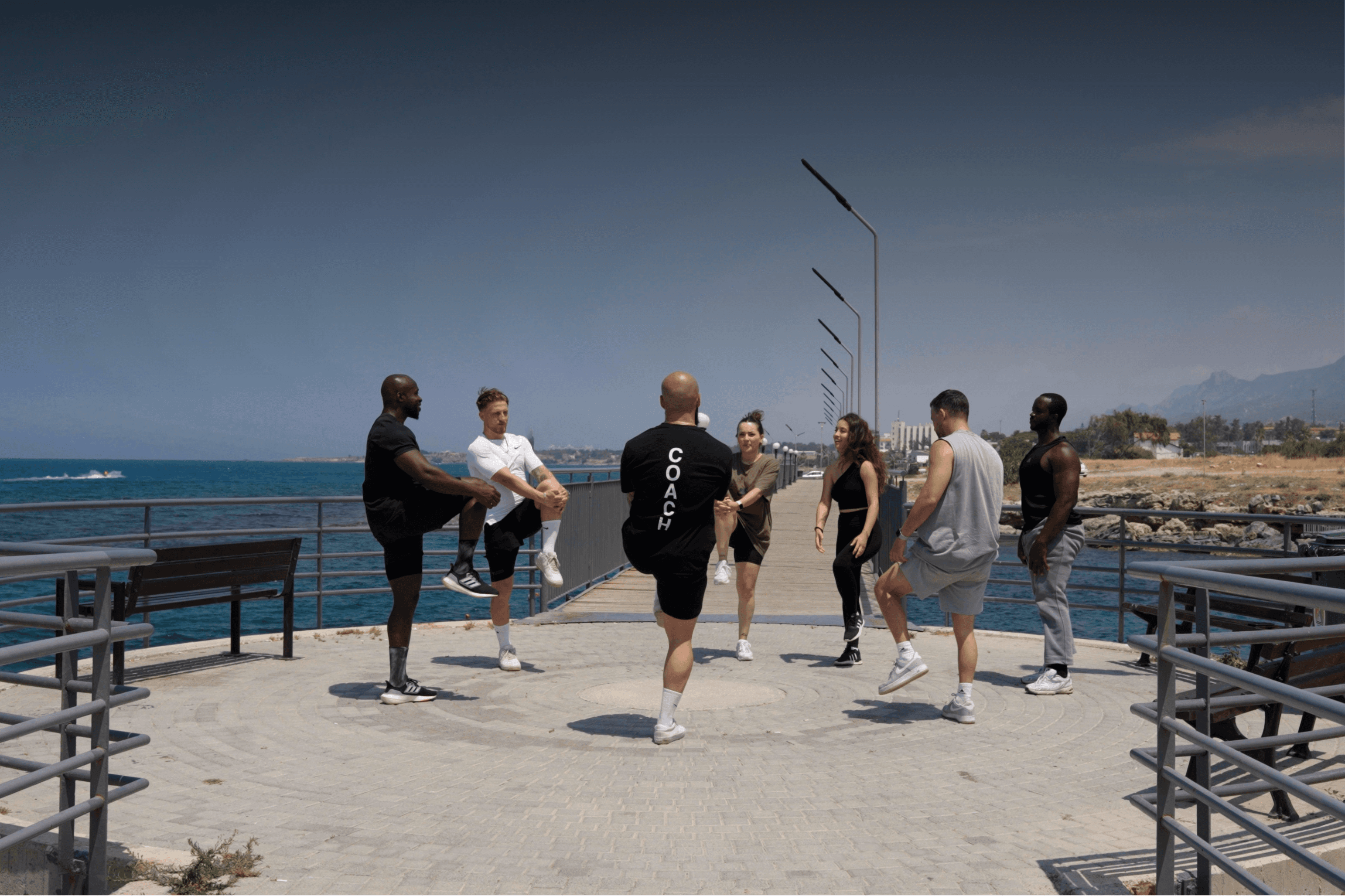 A group of people performing outdoor leg stretches on a sunny pier led by a fitness coach. Text reads: RETREAT with a Learn More button
