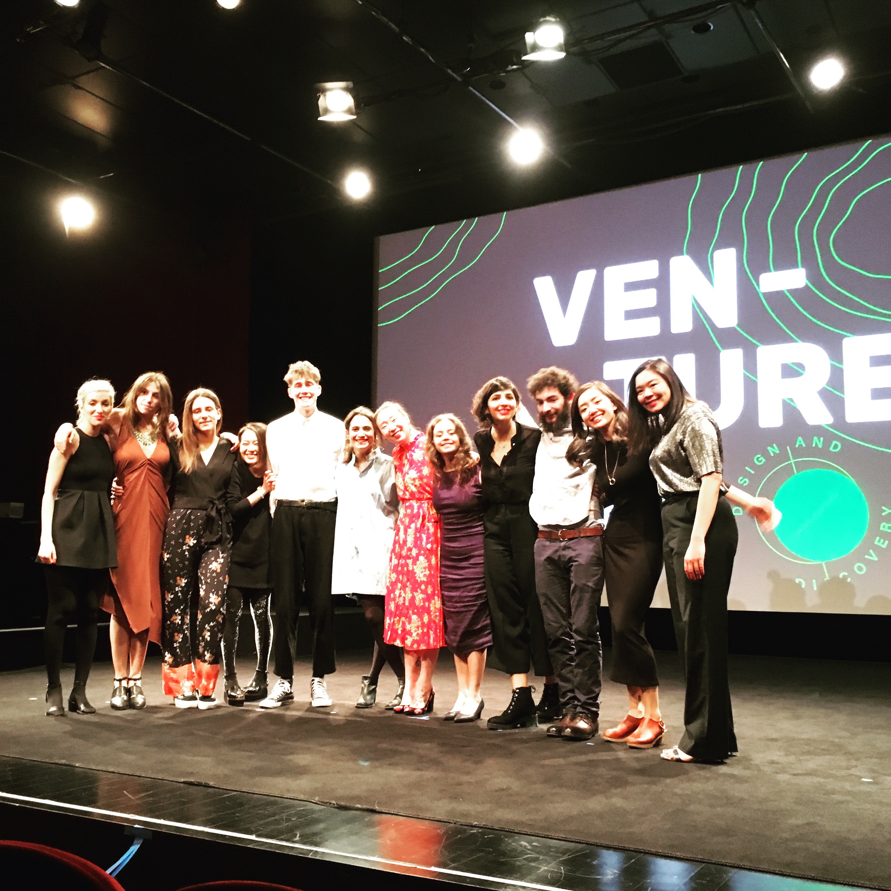 A photo of a group of people sitting on a stage in front of a screen projector that has on it the text: Ven-Ture.