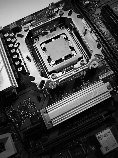 Czarno-białe zdjęcie z bliska przedstawiające procesor AMD Ryzen osadzony w gnieździe AM5 na płycie głównej Gigabyte. Na środku procesora widoczna jest świeżo nałożona warstwa pasty termoprzewodzącej; w tle widoczny radiator sekcji zasilania oraz sloty pamięci RAM.