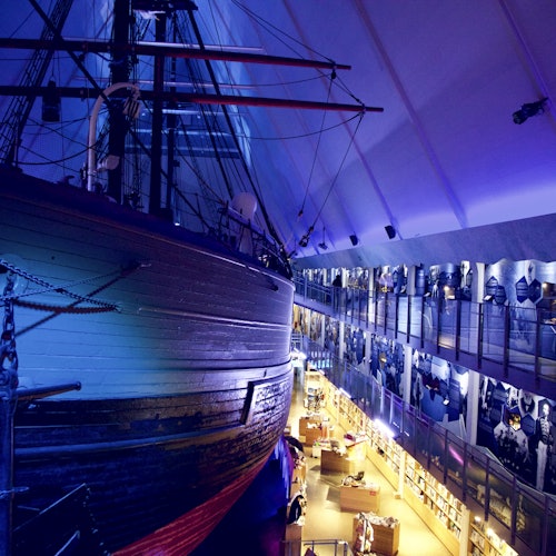 Большой деревянный корабль, выставленный в помещении под голубым освещением, рядом с двухуровневой галереей с историческими экспонатами и посетителями.