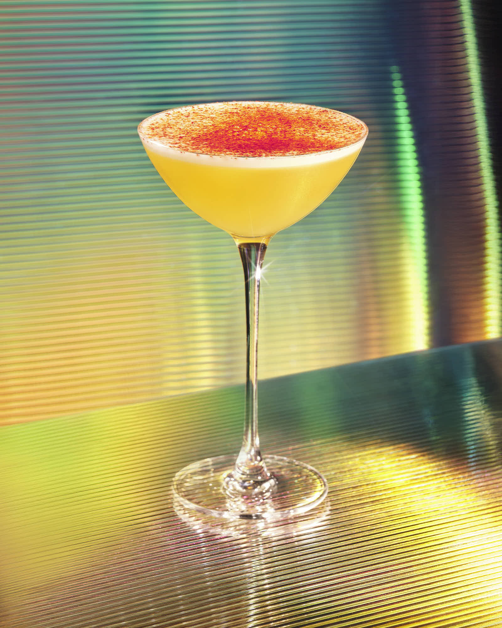 A vibrant cocktail in a tall glass, featuring a yellow drink topped with a red garnish, set against a colorful background.