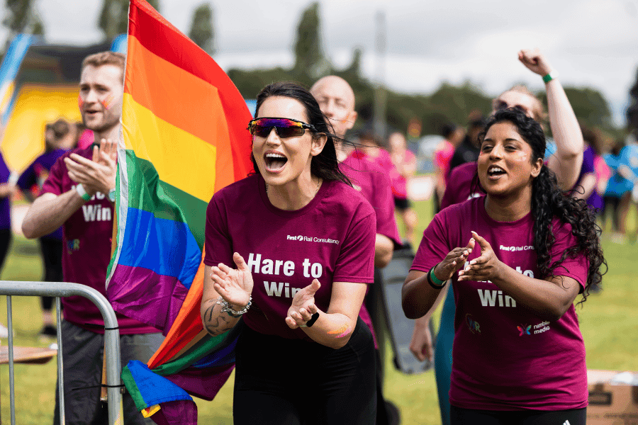 Outdoor team-building event with participants wearing matching blue shirts, taking part in inflatable obstacle courses and games, smiling and cheering, with the words 'ENTER A TEAM' in large bold white and green text across the image.