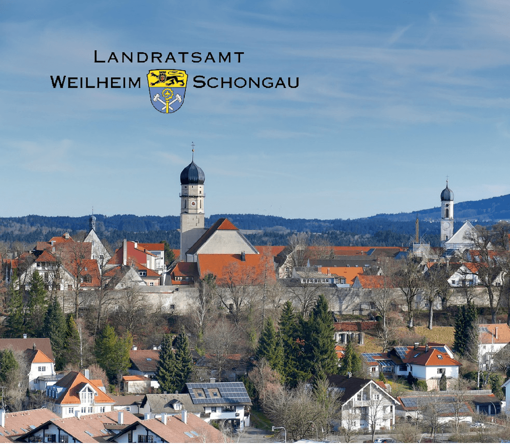 Eine Drohnenaufnahme des Ortes Weilheim in Schongau, darauf sind Gebäude sowie Kirchen des Ortes zu sehen, im Hintergrund erstreckt sich ein Bergpanorama, darauf befindet sich das Logo des Lanratsamt Weilheim Schongau