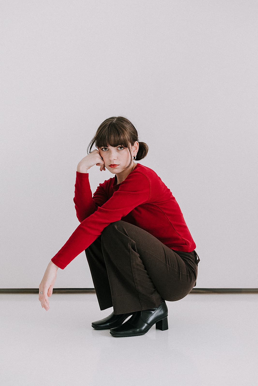 A model squatting in a red top and brown pants, with a pensive expression, part of a 90s-themed photoshoot at Revelator Studio, a Shreveport natural light studio.