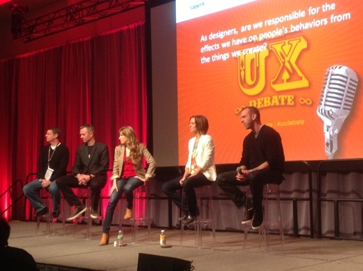 A photo of a group of people sitting on a stage in front of a screen projector. On it there is an orange banner with yellow letters that say: UX.