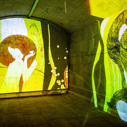 Sala de pedra arqueada iluminada com projeções coloridas de arte abstrata e floral, apresentando uma figura com cabelos esvoaçantes.