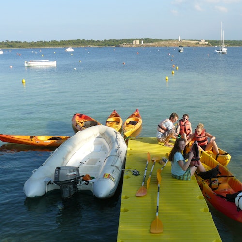 People sitting on a yellow dock by the ocean, surrounded by kayaks and an inflatable boat, with buoys and sailboats in the distance.