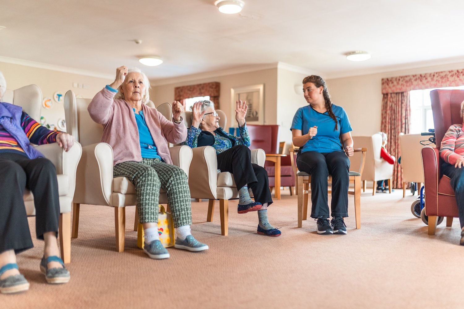 Group of elderly people seated in a circle participating in a light exercise session led by a caregiver.