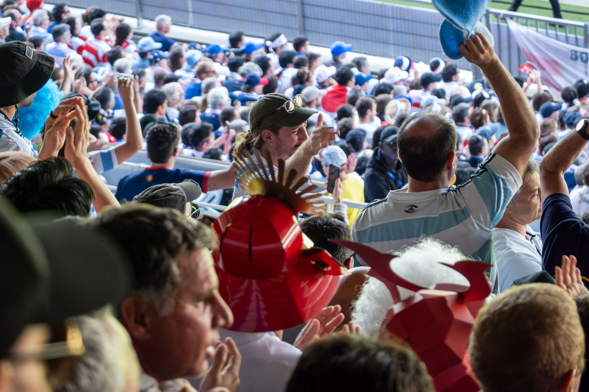Tribune qui applaudit, casques rouges en carton à crête dorée au premier plan, un supporter en maillot argentin brandit un bonnet bleu
