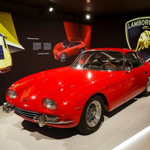 Coche vintage rojo en exhibición en un entorno similar a un museo con la marca Lamborghini y otras imágenes de coches en la pared detrás.