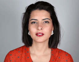 A woman with short, dark hair wears a red top with small blue dots. She has dark eyebrows, dark eyes, and is wearing small silver earrings. Her lips are painted red, and she has a neutral expression. The background is a solid light gray.