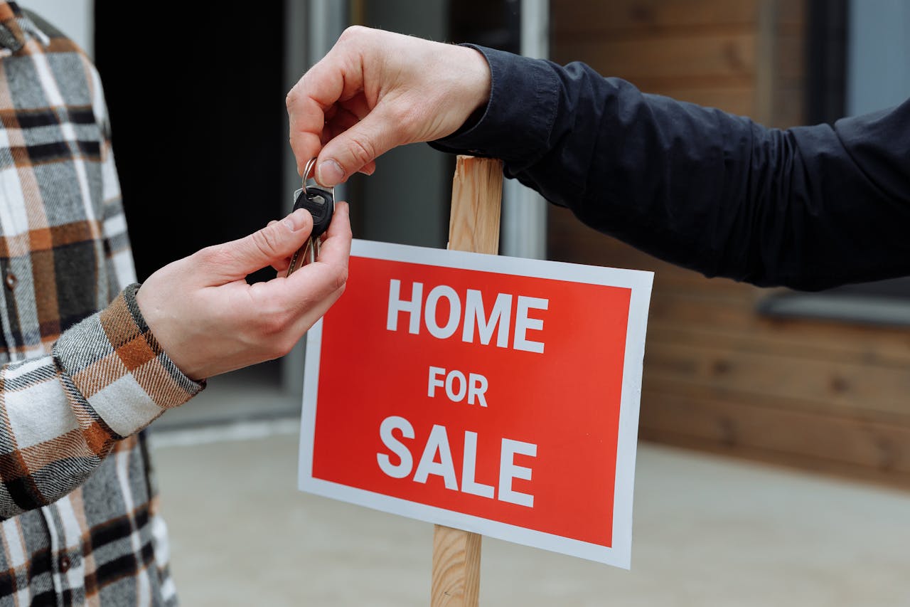 Image that shows signals the sell of a home. A hand giving another hand a key, in front a signal that "Home for Sale".