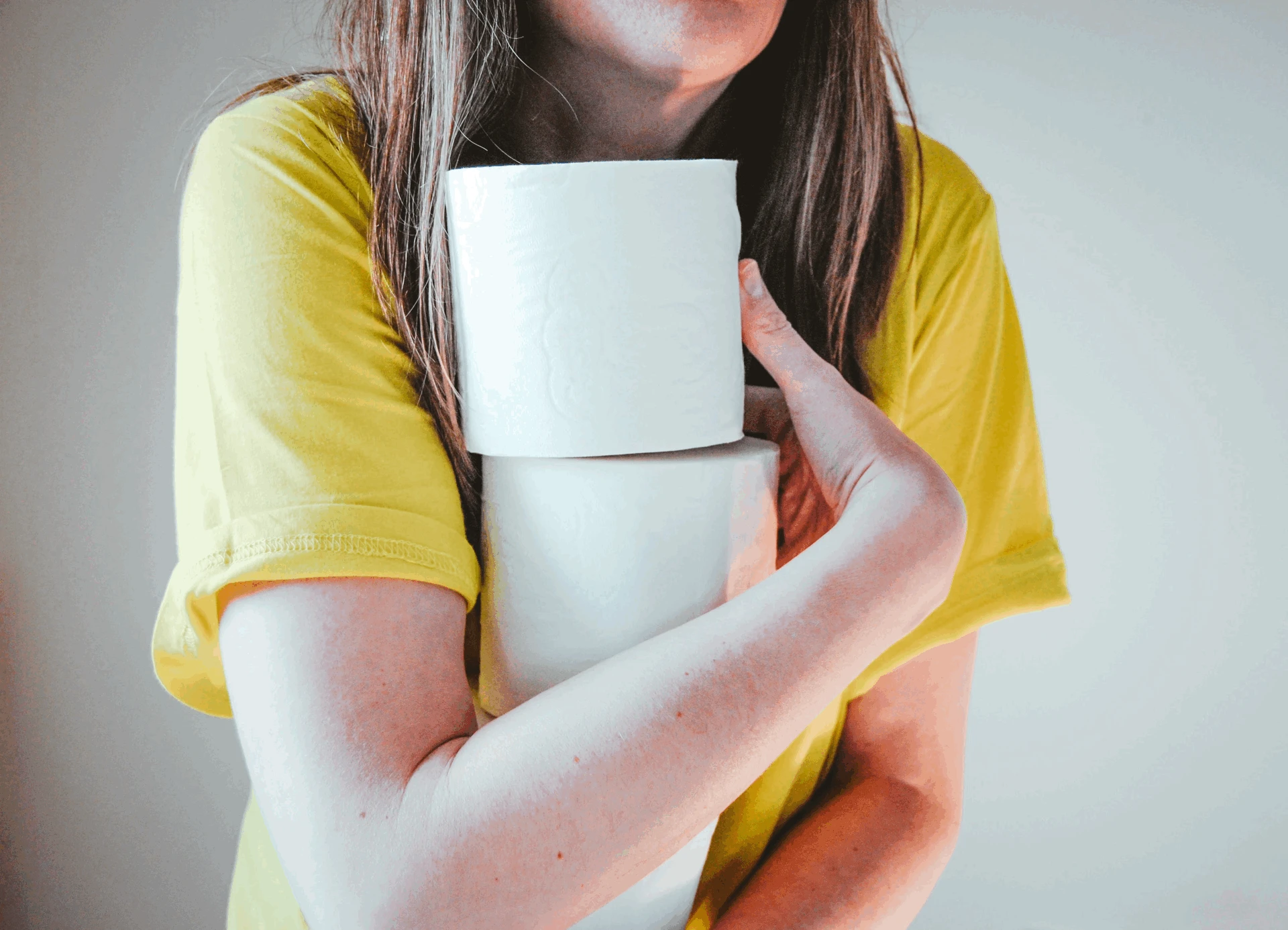 Woman in a yellow shirt holding two large rolls of toilet paper against her chest.